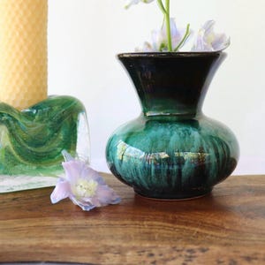 May include: A small ceramic vase with a glossy finish featuring a deep emerald green and black drip glaze pattern. The vase has a rounded body and a flared rim, containing light purple flowers with white centres, positioned near a candle.