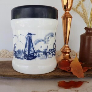 May include: A white glass canister with a dark blue lid and a blue nautical illustration featuring sailboats and a windmill. The cylindrical container has a smooth finish and a repeating maritime scene printed around the exterior surface.