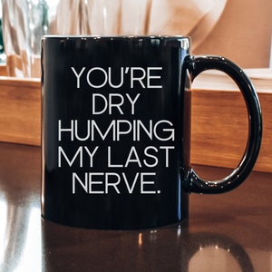 May include: A black ceramic mug with the text "YOU'RE DRY HUMPING MY LAST NERVE" in white. The mug has a curved handle and is sitting on a wooden surface. The mug is part of a kitchenware collection.