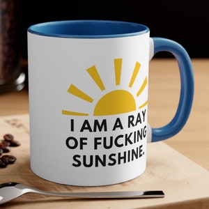 May include: White coffee mug with a blue rim and handle. The mug has a yellow sun graphic and the text "I am a ray of fucking sunshine."