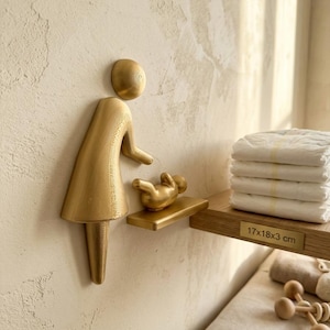 May include: A gold-colored wall-mounted sign depicting a stylized figure tending to a baby on a small shelf. The sign features a smooth, rounded head, a dress-like body, and a small infant figure resting on a horizontal platform. The shelf has a label reading 17x18x3 cm.
