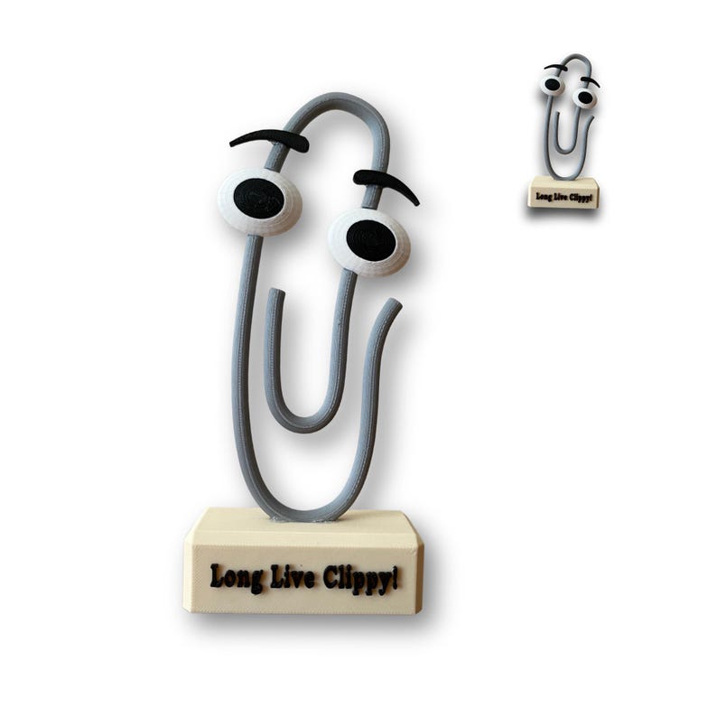 May include: A grey paperclip with googly eyes and black eyebrows is standing on a small wooden base. The base has the words "Long Live Clippy!" printed on it.