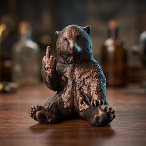 Op de afbeelding: Een bronzen beerbeeldje, zittend met de middelvinger omhoog. De beer heeft een getextureerd oppervlak en staat tegen een vage achtergrond met flessen en gereedschap, wat duidt op een werkplaats of studio.