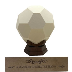 Puede incluir: Una esfera geométrica blanca sobre una base de madera con el texto "A NEW HAND TOUCHES THE BEACON" en una tarjeta de papel marrón.