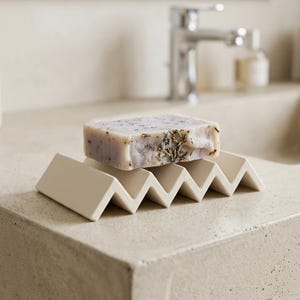 Puede incluir: Una pastilla de jabón rectangular con hierbas visibles descansa sobre un jabonera crema en forma de zigzag. El jabón tiene un tono morado claro con motas verdes y marrones. La jabonera es de cerámica y está sobre una encimera clara.