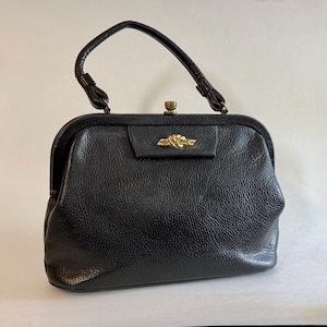 May include: A black handbag with a textured surface, featuring a single top handle and a gold-toned metal clasp closure. The front of the bag is adorned with a small, horizontal gold-toned decorative element shaped like a knot.