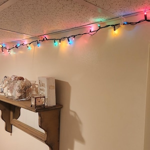 May include: A string of colorful Christmas lights hangs from the ceiling of a room with white walls. The lights are multi-colored, including red, green, blue, yellow, and pink. There is a wooden shelf on the wall with decorative items on it.