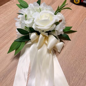 May include: A floral arrangement featuring white artificial roses and hydrangea blossoms accented with green leaves. The bouquet is tied with long, flowing cream-coloured satin ribbons that drape downwards from the base of the flower cluster.