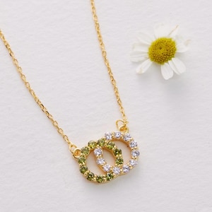 May include: A gold necklace featuring two intertwined circles, one set with green gems and the other with clear crystals. A small white daisy with a yellow centre lies on a white surface nearby.