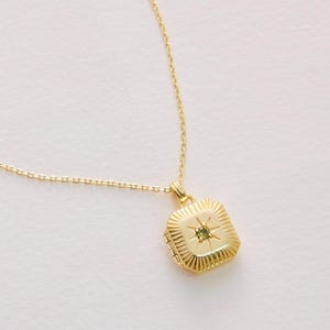 May include: A gold-colored necklace with a square pendant. The pendant has a starburst design and a small gemstone in the center. The necklace is on a white background.