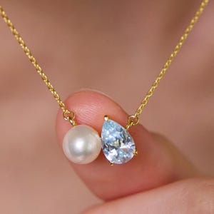Pode incluir: Um delicado colar de ouro com uma pérola e um pingente de pedra preciosa azul claro em forma de gota. O colar é segurado pelos dedos de uma pessoa, mostrando os detalhes finos da joia.