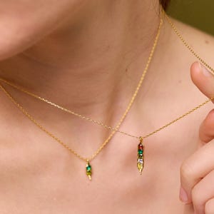 May include: Two gold necklaces with delicate chains. One necklace features a pendant with a green gem, while the other has a pendant with a row of colorful gems. The necklaces are worn around the neck.