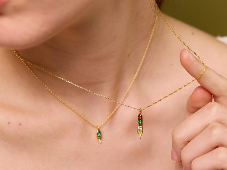 May include: Two gold necklaces with delicate chains. One necklace features a pendant with a green gem, while the other has a pendant with a row of colorful gems. The necklaces are worn around the neck.