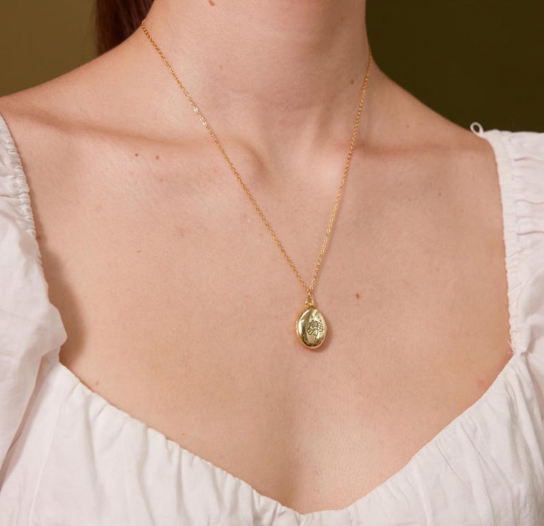 Puede incluir: Un collar de cadena dorada con un peque&ntilde;o colgante de medall&oacute;n ovalado. El medall&oacute;n tiene un dise&ntilde;o floral.