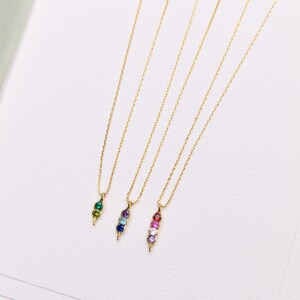 May include: Four gold-colored necklaces with pendant charms. Each charm is a pod shape, filled with colorful gemstones. The gemstones are green, blue, and pink. The necklaces are on a white surface.