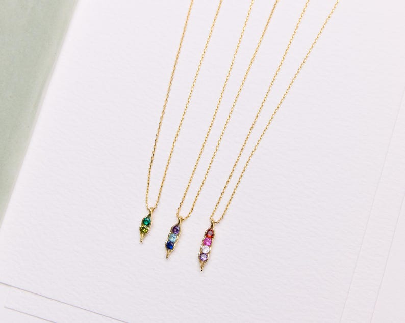 May include: Four gold-colored necklaces with pendant charms. Each charm is a pod shape, filled with colorful gemstones. The gemstones are green, blue, and pink. The necklaces are on a white surface.