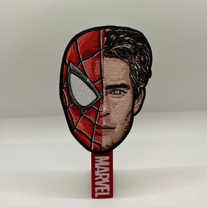 May include: A red and black embroidered patch of Spider-Man's face with the right side of his face showing Tom Holland's face. The patch is attached to a red backing with the word "MARVEL" in white lettering.