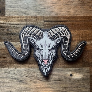 May include: An embroidered patch featuring the head of a ram with large, curved horns. The design uses black, white, and grey thread with a small pink accent on the nose. The ram is depicted facing forward with detailed shading and a black border.