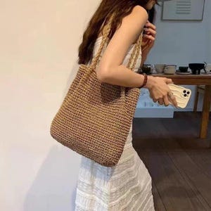 Könnte beinhalten: Eine gewebte Tragetasche in Naturtönen. Die Tasche hat eine rechteckige Form mit geflochtenen Henkeln. Die Textur der Tasche scheint aus einem strohähnlichen Material zu bestehen, das sich zum Tragen von Gegenständen eignet.