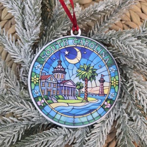 May include: A round, flat ornament featuring a stained-glass style illustration of South Carolina. The design includes a state house building, a palmetto tree, a lighthouse, and a crescent moon under the text SOUTH CAROLINA, suspended by a red ribbon.