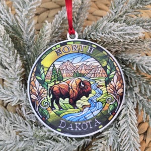 May include: A round, flat ornament featuring a stained-glass style illustration of a bison standing near a stream with mountains in the background. The text NORTH DAKOTA is printed in a circular border. A red ribbon is attached to the top.