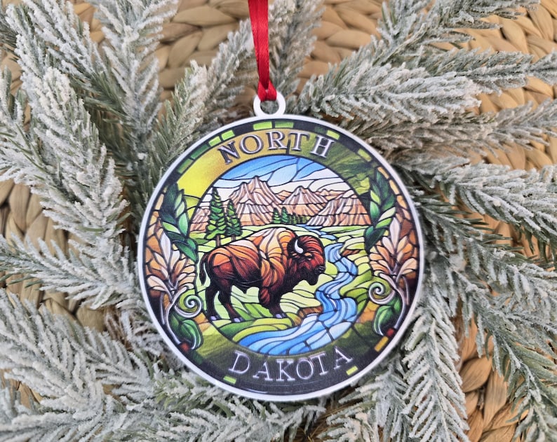 May include: A round, flat ornament featuring a stained-glass style illustration of a bison standing near a stream with mountains in the background. The text NORTH DAKOTA is printed in a circular border. A red ribbon is attached to the top.