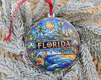 May include: A round Christmas ornament with a Florida theme. The ornament features a stained-glass design with a beach scene, palm trees, a lighthouse, and the word "FLORIDA". It has a red ribbon for hanging.