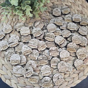 May include: A collection of small, circular wooden tokens, each engraved with a unique illustration and the name of a U.S. National Park, including labels like Zion, Yosemite, Yellowstone, Denali, and Gateway Arch, arranged in a scattered pile.