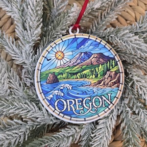 May include: A round, flat ornament featuring a stained-glass style illustration of an Oregon coastal landscape with a mountain, forest, ocean waves, and a sun. The word OREGON is printed in white capital letters at the bottom of the circular design.
