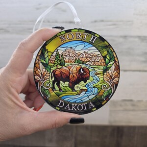 May include: A round, stained-glass style ornament featuring a brown bison standing in a green landscape with a blue river, pine trees, and mountains. The text NORTH DAKOTA is printed in white along the top and bottom edges of the circular frame.