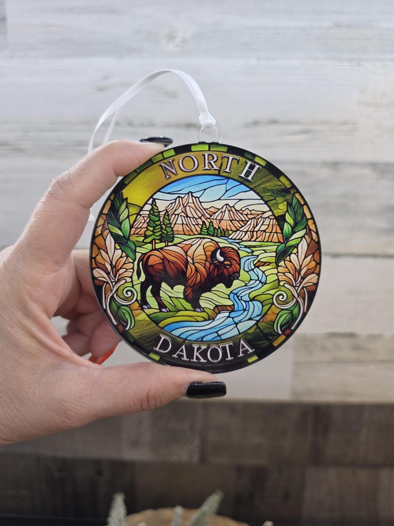May include: A round, stained-glass style ornament featuring a brown bison standing in a green landscape with a blue river, pine trees, and mountains. The text NORTH DAKOTA is printed in white along the top and bottom edges of the circular frame.