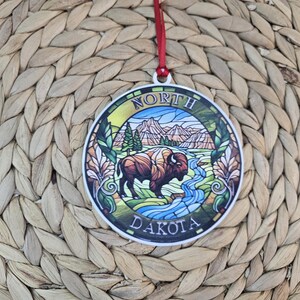 May include: A round, flat ornament featuring a stained-glass style illustration of a bison standing near a blue river with mountains and pine trees in the background. The text NORTH DAKOTA is printed in white along the circular border.