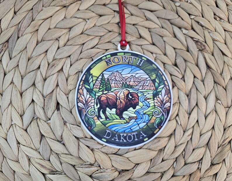 May include: A round, flat ornament featuring a stained-glass style illustration of a bison standing near a blue river with mountains and pine trees in the background. The text NORTH DAKOTA is printed in white along the circular border.