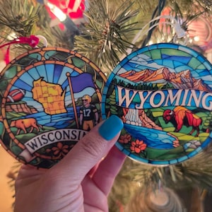 May include: Two circular ornaments with a stained glass effect. The left ornament features a map of Wisconsin, a football, a cow, and a football player with the text WISCONSIN. The right ornament depicts a mountain landscape, a waterfall, and a bison with the text WYOMING.