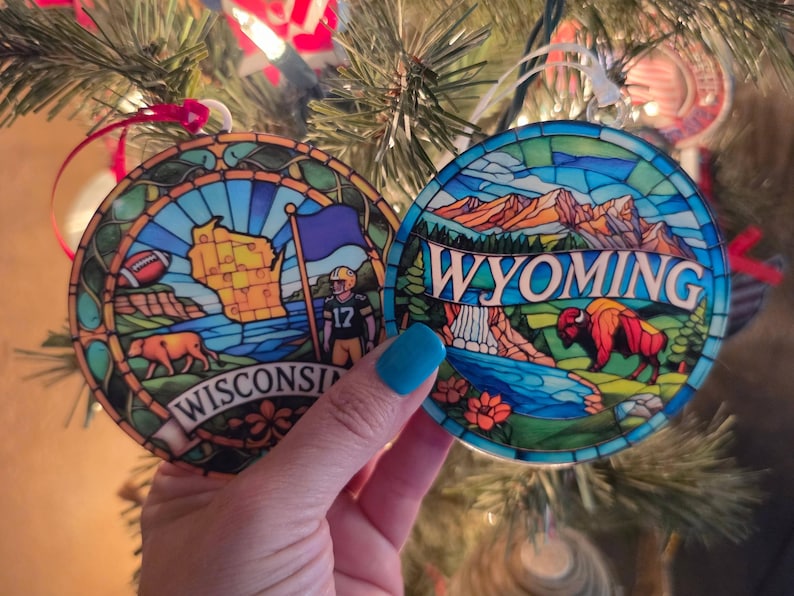 May include: Two circular ornaments with a stained glass effect. The left ornament features a map of Wisconsin, a football, a cow, and a football player with the text WISCONSIN. The right ornament depicts a mountain landscape, a waterfall, and a bison with the text WYOMING.