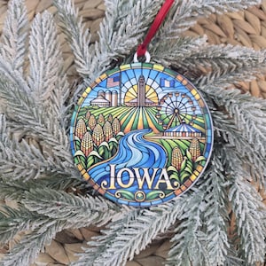 May include: A round, stained-glass style ornament featuring a colorful illustration of Iowa. The design includes corn stalks, a winding blue river, a Ferris wheel, and the Iowa State Capitol building. The word IOWA is printed in large, bold letters.