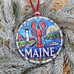 May include: A round, stained-glass style ornament featuring a red lobster, a white lighthouse, a boat, and a sun over a coastal landscape. The word MAINE is printed in large, bold, white capital letters at the bottom of the circular design.