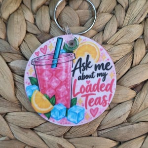 May include: A round keychain featuring a graphic of a pink iced drink in a clear cup with a blue straw, lemon slices, and ice cubes. The text reads Ask me about my Loaded Teas! in black and pink script, accented with small yellow stars and pink hearts.