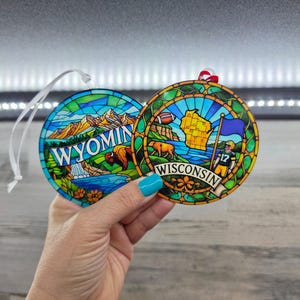 May include: Two round ornaments featuring stained glass style designs. The left ornament displays a mountain landscape with a bison and the text WYOMING. The right ornament shows a map of Wisconsin, a football player, and the text WISCONSIN.