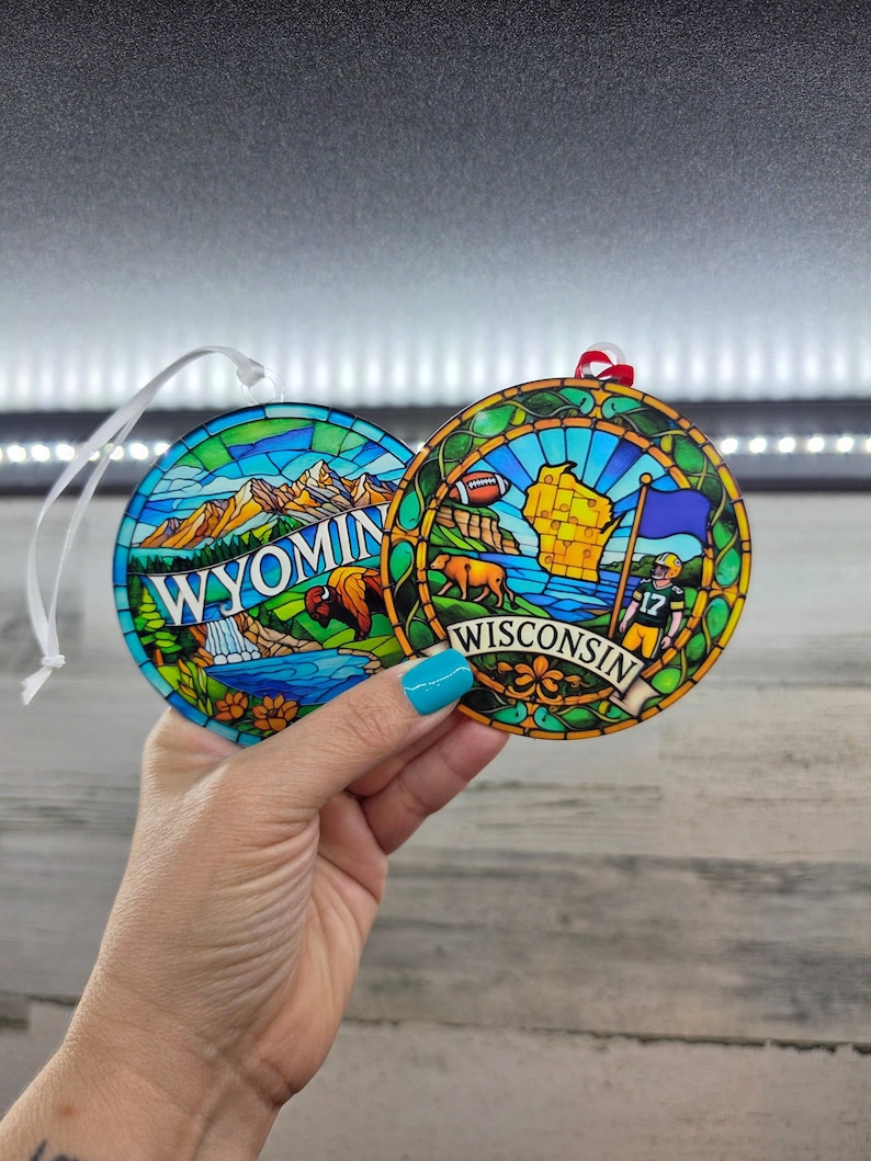 May include: Two round ornaments featuring stained glass style designs. The left ornament displays a mountain landscape with a bison and the text WYOMING. The right ornament shows a map of Wisconsin, a football player, and the text WISCONSIN.