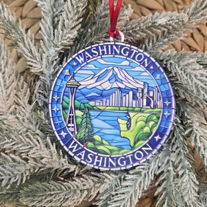 May include: A circular ornament featuring a stained-glass style illustration of the Washington landscape, including the Space Needle, a mountain, and a city skyline. The word WASHINGTON is printed twice along the blue border with a red ribbon.