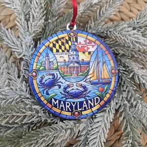 May include: A round, stained-glass style ornament featuring Maryland imagery, including the state flag, the Maryland State House, a sailboat, and two blue crabs. The word MARYLAND is printed in bold letters at the bottom, and it hangs from a red ribbon.