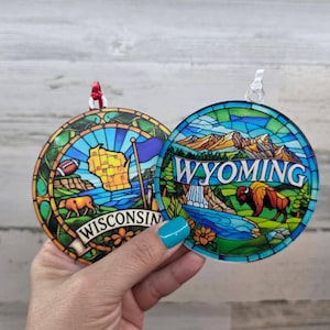 May include: Two round ornaments featuring stained glass style designs. One displays a map of Wisconsin with a football and the text WISCONSIN. The other shows a Wyoming mountain landscape with a bison and the text WYOMING.