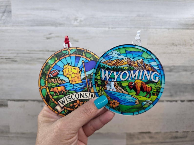 May include: Two round ornaments featuring stained glass style designs. One displays a map of Wisconsin with a football and the text WISCONSIN. The other shows a Wyoming mountain landscape with a bison and the text WYOMING.