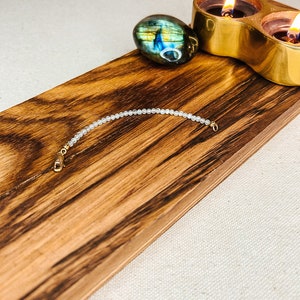 May include: A delicate white beaded bracelet with gold clasps, displayed on a wooden surface with a labradorite crystal and a gold candle holder.