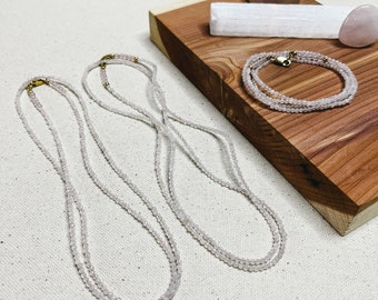 May include: Three delicate rose quartz necklaces with gold clasps, displayed on a wooden surface with a white candle and a selenite wand.