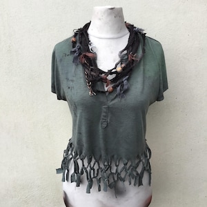 May include: A green short-sleeved shirt with a fringed hemline and a decorative necklace. The necklace is made of dark fabric, wooden beads, and metal accents. The shirt is displayed on a mannequin.