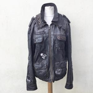 Peut inclure: Veste en cuir noir vieilli avec plusieurs poches, des écussons et un emblème de tête de mort et d'os croisés. La veste est dotée d'une fermeture éclair sur le devant, d'un col avec des œillets métalliques et de coutures décoratives. La veste est présentée sur un mannequin.