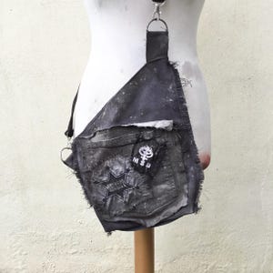 May include: A black denim crossbody bag with a distinctive, asymmetrical shape. It has a large pocket with a star and skull patch, and the letters "MSU". The bag has a raw, frayed edge and a silver chain strap.