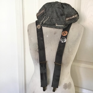 May include: A grey tweed newsboy cap with the word "DANGER" and chain details. The cap is on a mannequin with black suspenders, decorated with pins showing a biohazard symbol and a skull.